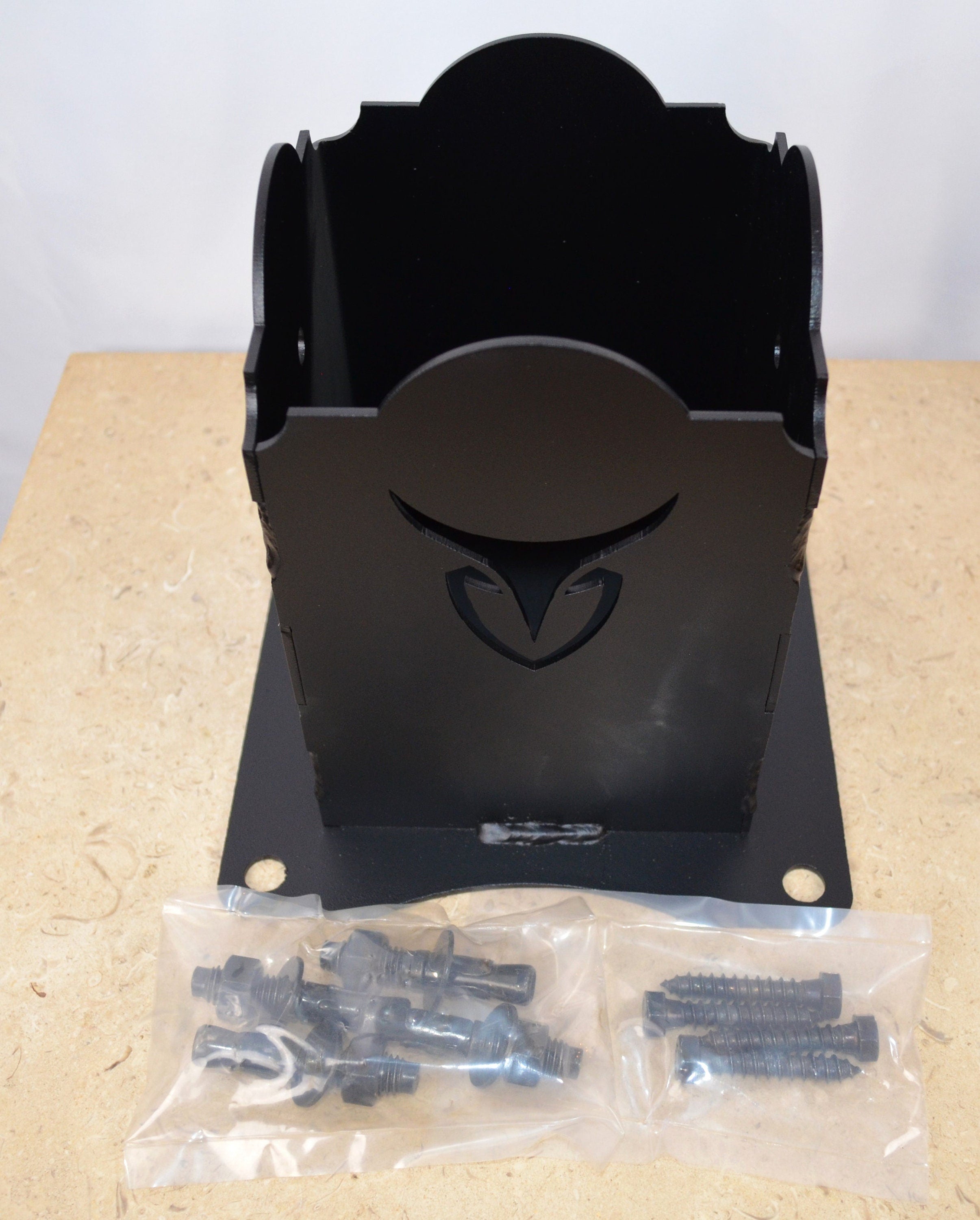 A stylish wooden post holder is sitting on a table, displaying a distinct design at its base. Laying in front of this bracket are bags with 4 concrete anchors and 4 lag screws.