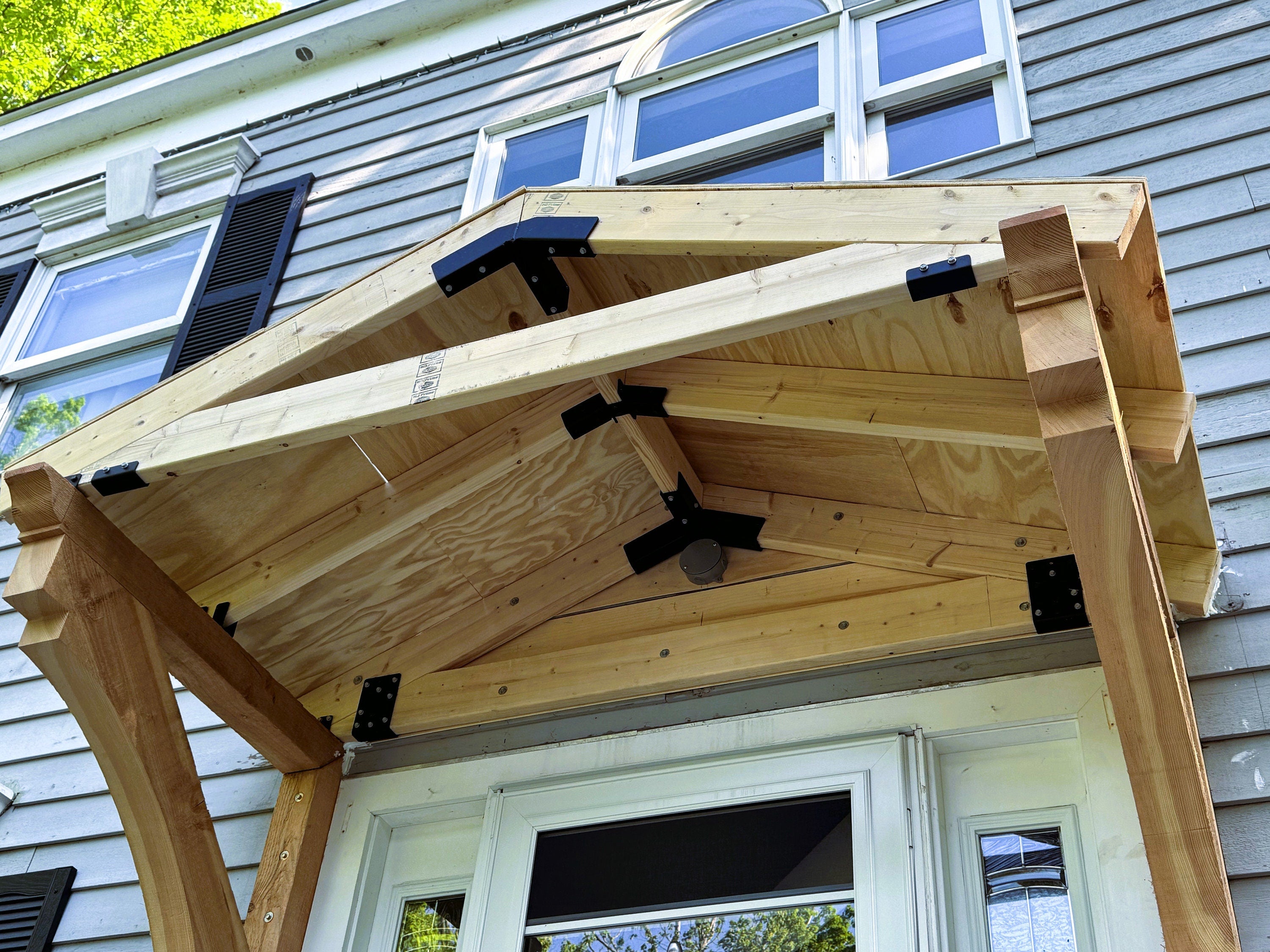 A newly constructed wooden porch roof showcases its triangular design and robust support beams, enhancing the entryway of a home on a sunny day.