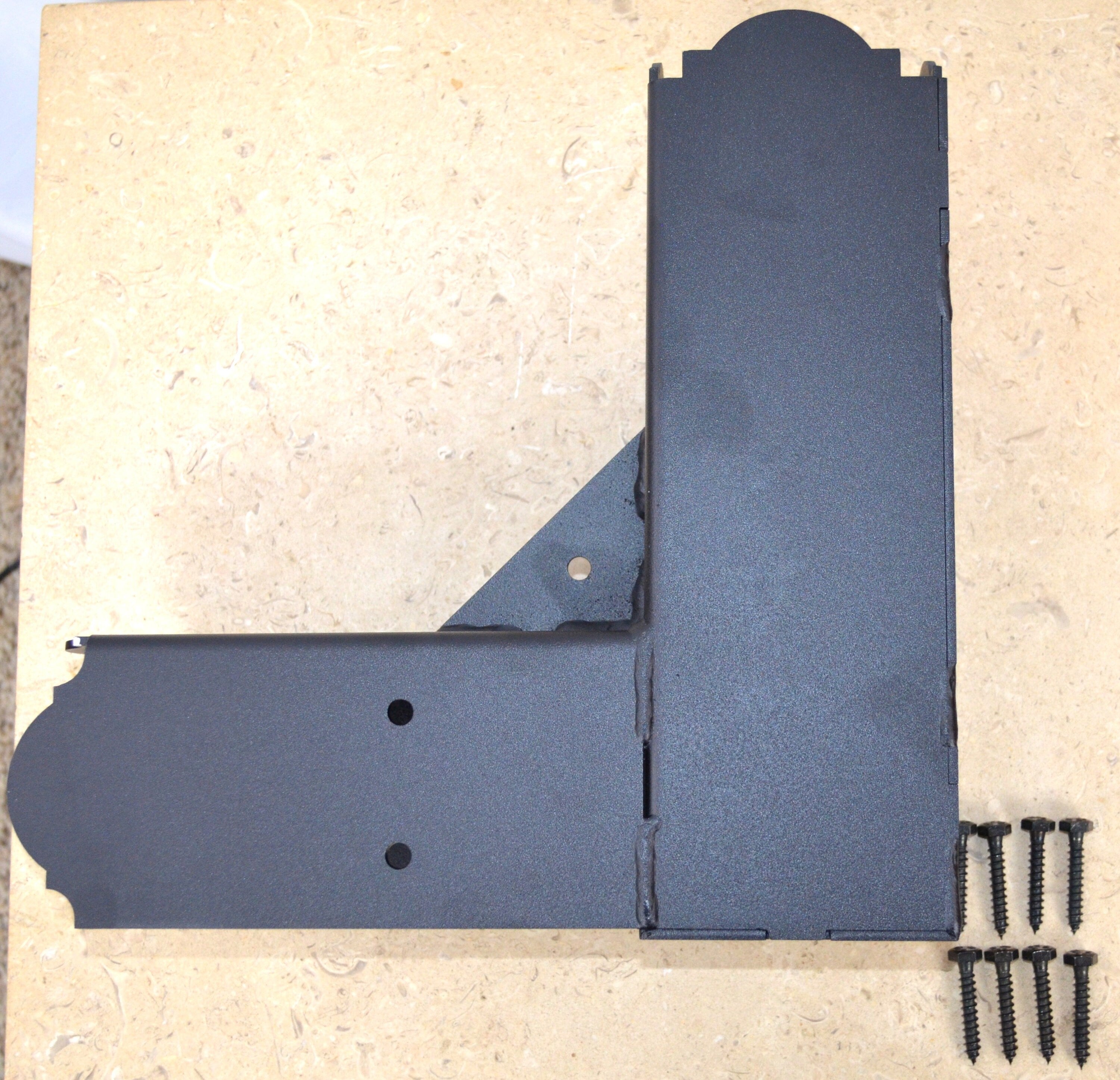 A black metal elbow bracket rests on a wooden tabletop alongside several screws, showcasing its design for assembly or construction projects in a workshop setting.