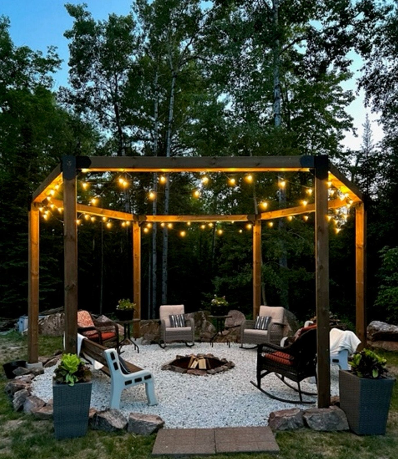 a 6x6 hexagon pergola with chairs and firepit, lit with string lights around the top edges in a forested setting