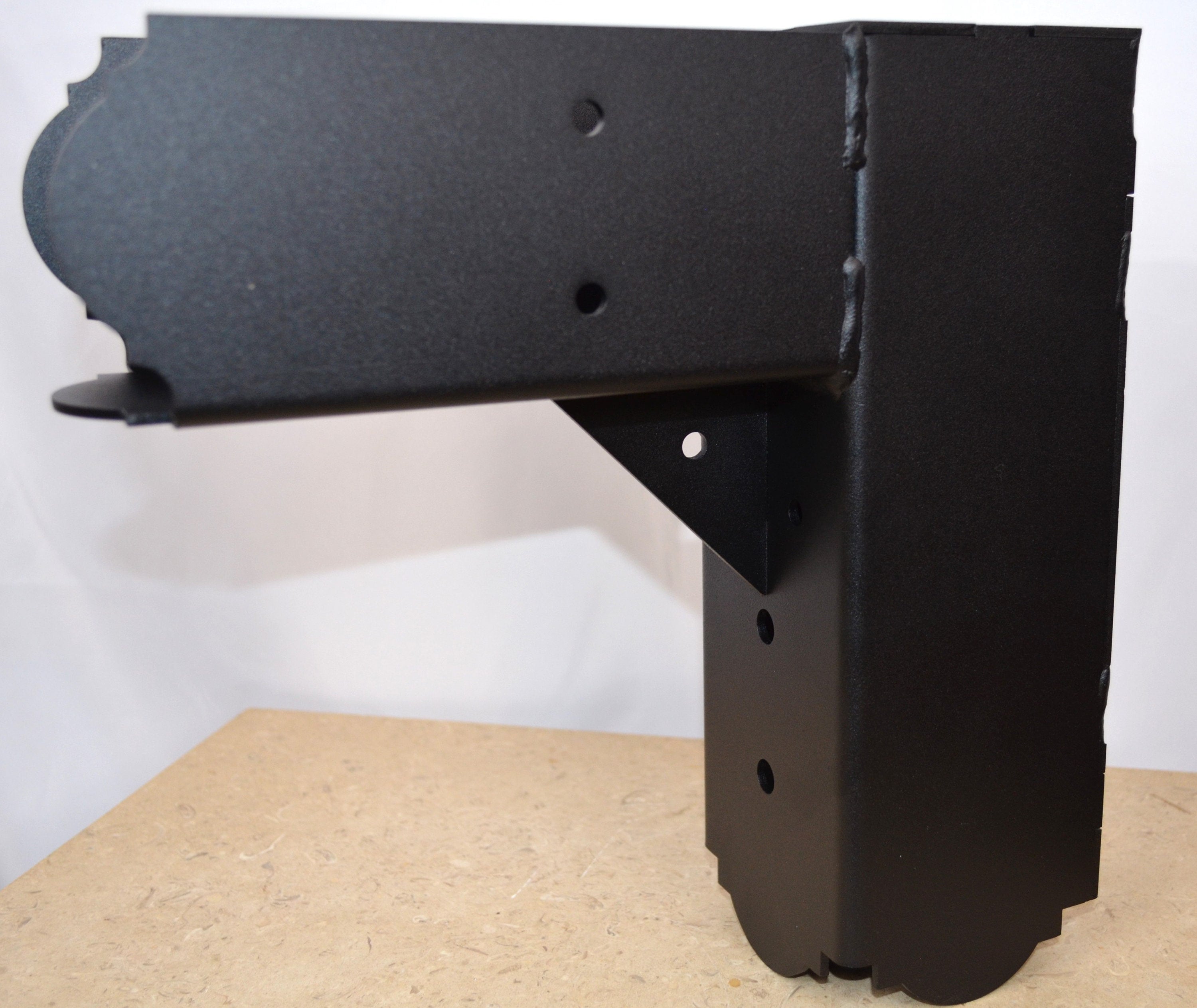 A black metal elbow bracket rests on a wooden tabletop alongside several screws, showcasing its design for assembly or construction projects in a workshop setting.