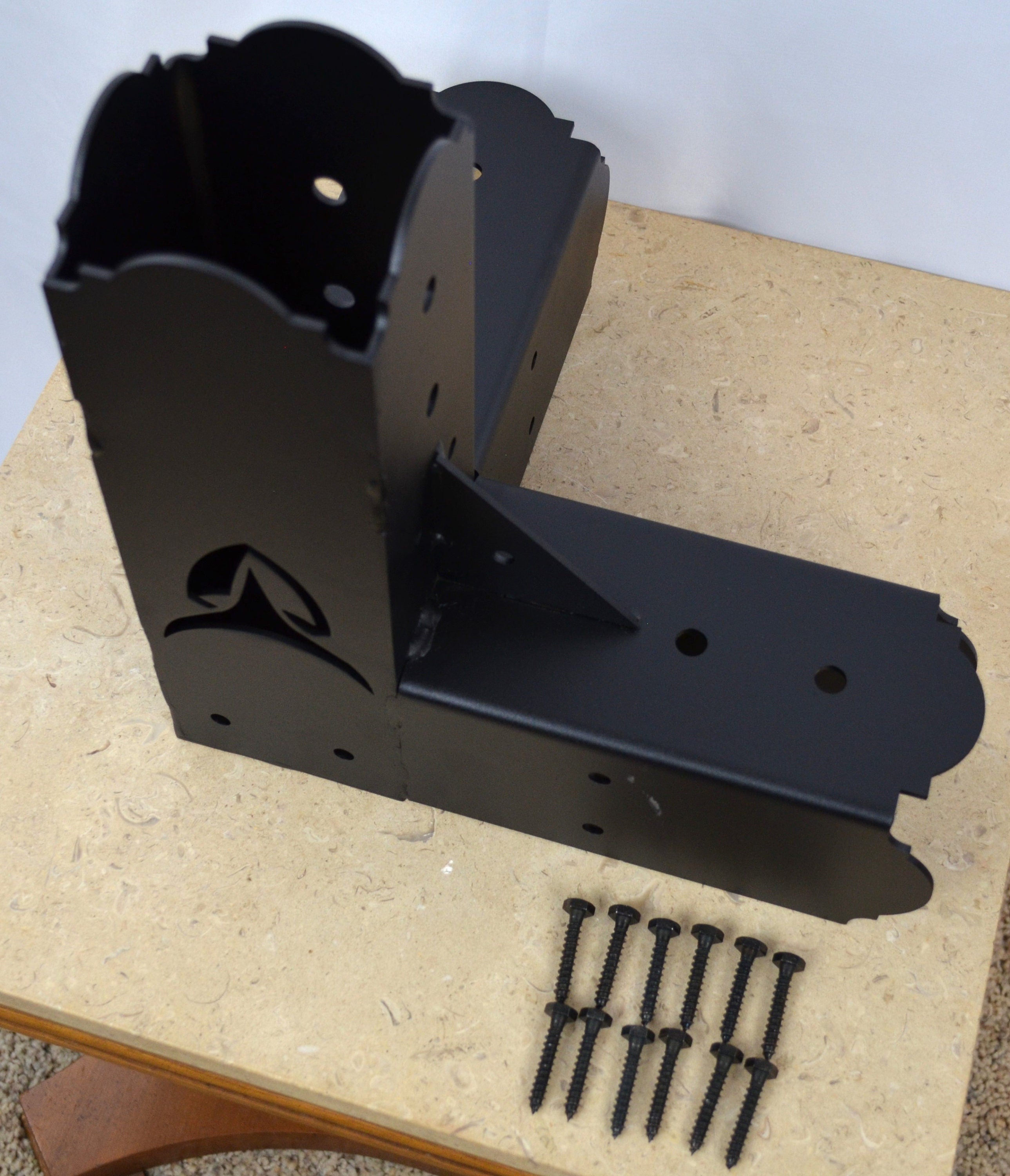 A black metal bracket rests on a wooden tabletop alongside several screws, showcasing its design for assembly or construction projects in a workshop setting.