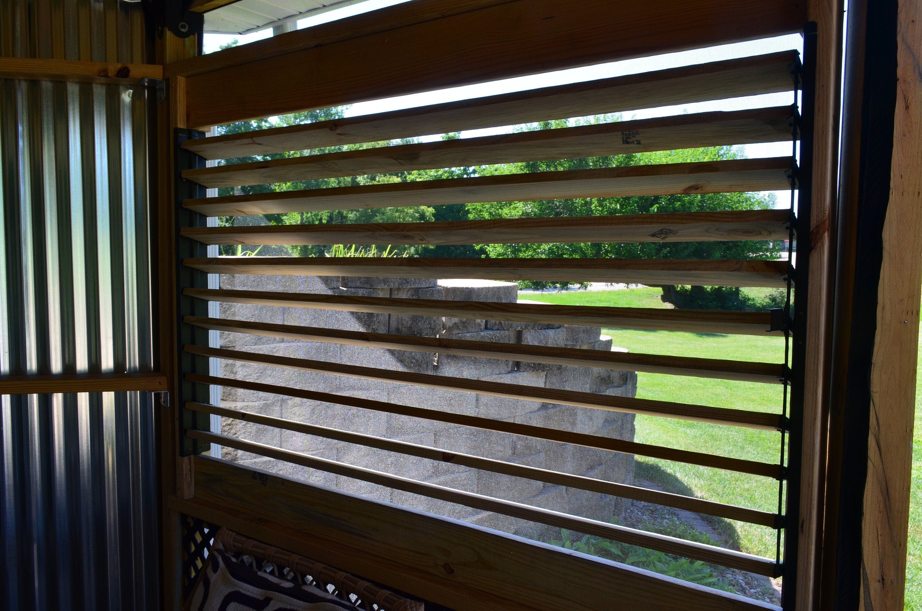 The view reveals a wooden slatted window framing a vibrant green landscape bathed in sunlight, inviting a sense of tranquility and connection with nature.