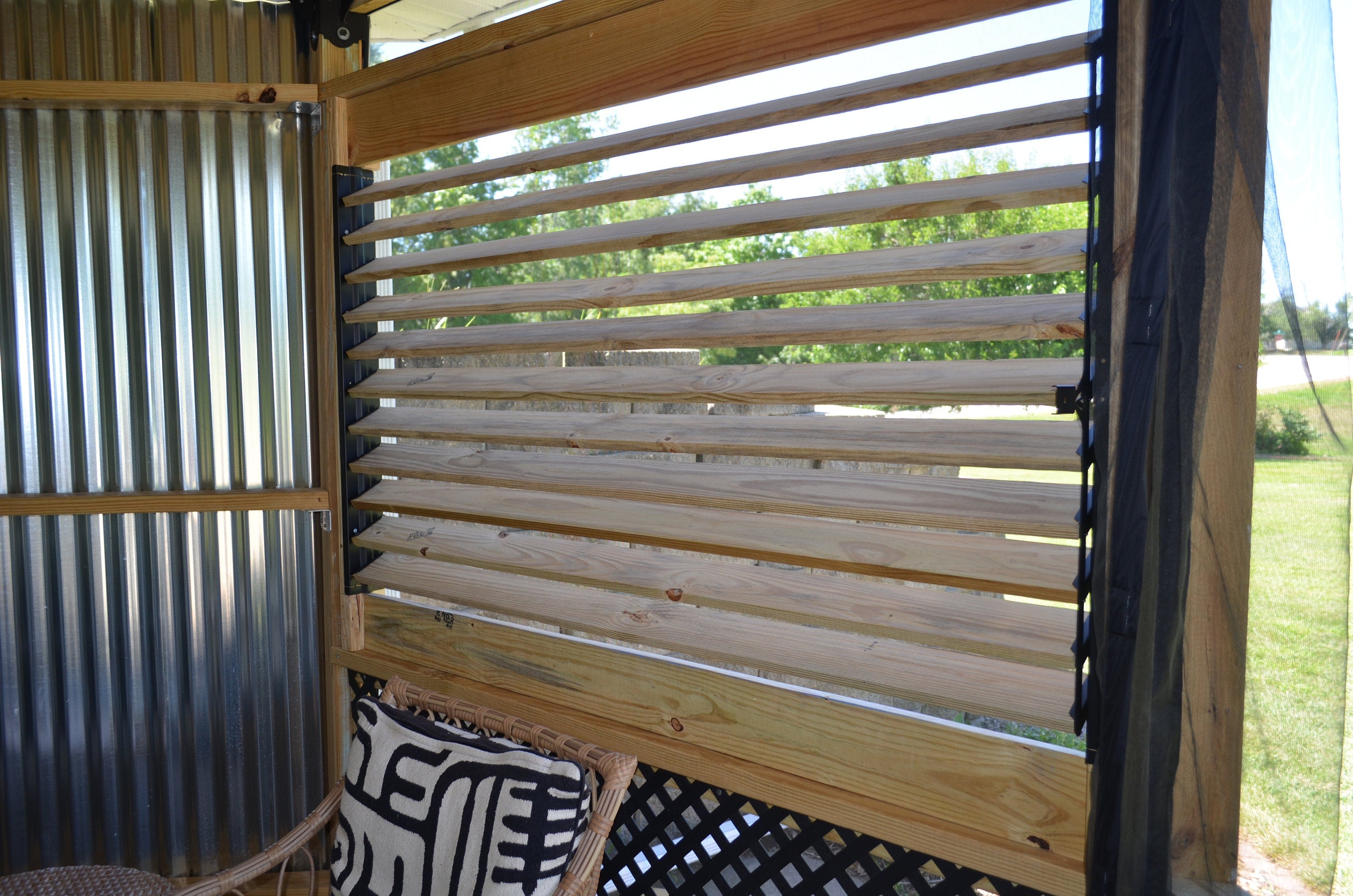 The view reveals a wooden slatted window framing a vibrant green landscape bathed in sunlight, inviting a sense of tranquility and connection with nature.