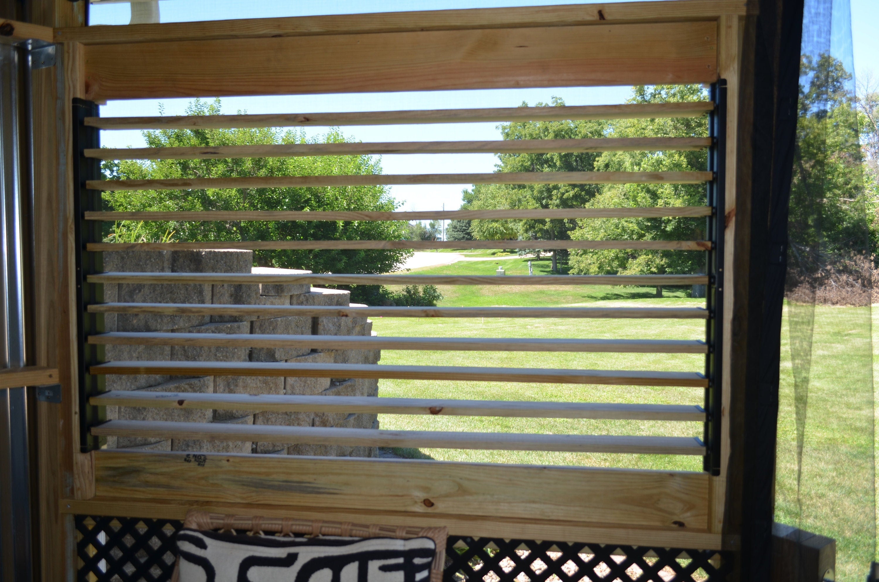 The view reveals a wooden slatted window framing a vibrant green landscape bathed in sunlight, inviting a sense of tranquility and connection with nature.