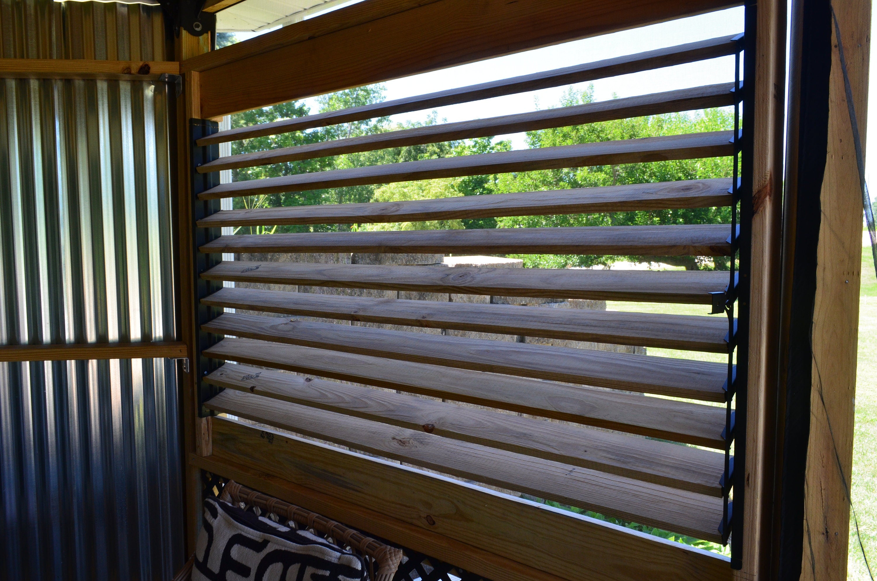 The view reveals a wooden slatted window framing a vibrant green landscape bathed in sunlight, inviting a sense of tranquility and connection with nature.