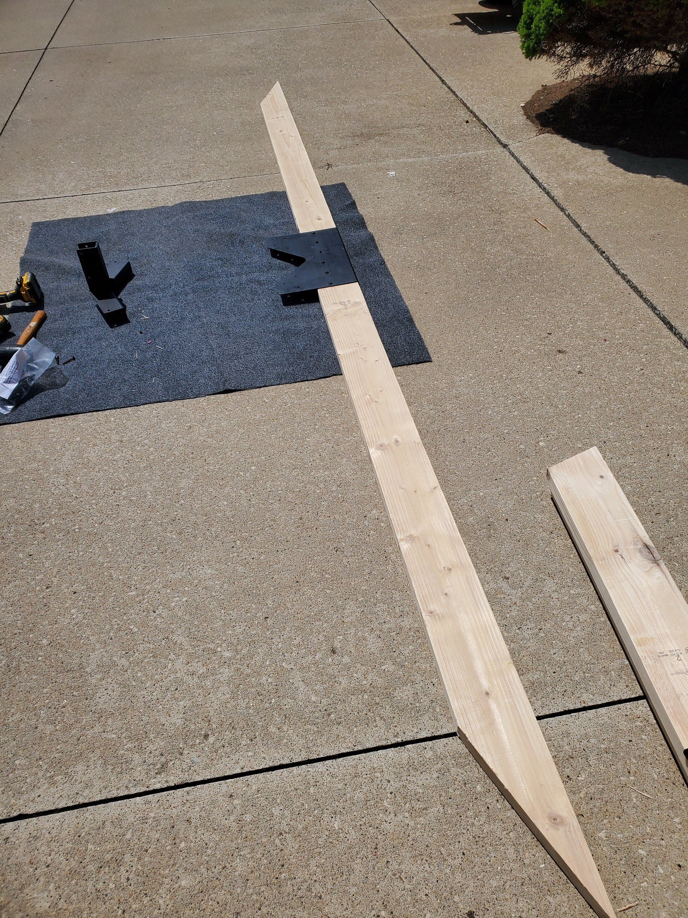 A wooden beam is positioned on a driveway, accompanied by tools and materials. The work is taking place in a suburban setting under bright sunlight, suggesting ongoing construction.