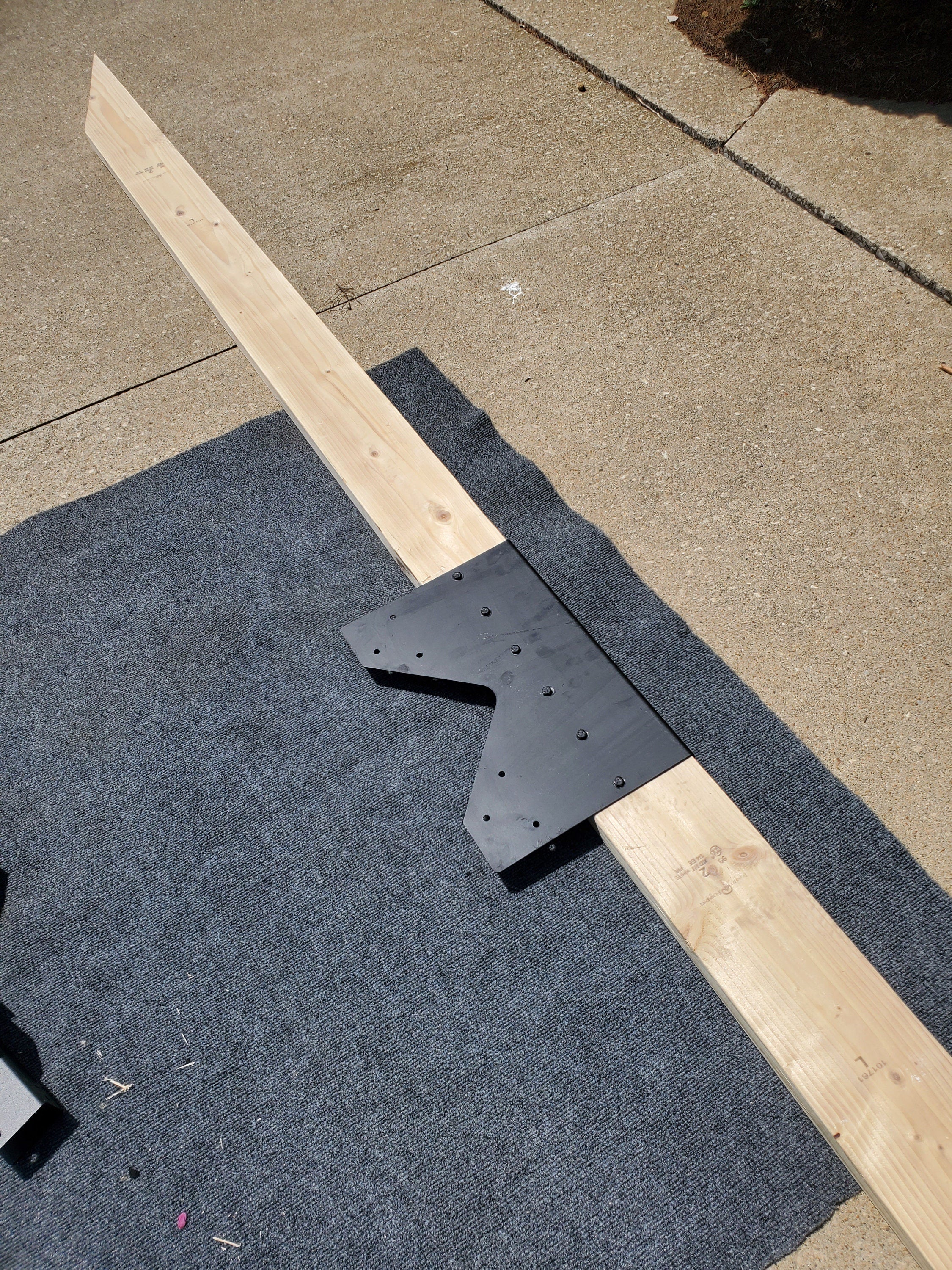 A wooden beam is positioned on a gray surface with a metal bracket attached, illustrating a DIY woodworking project underway in a residential driveway under bright sunlight.