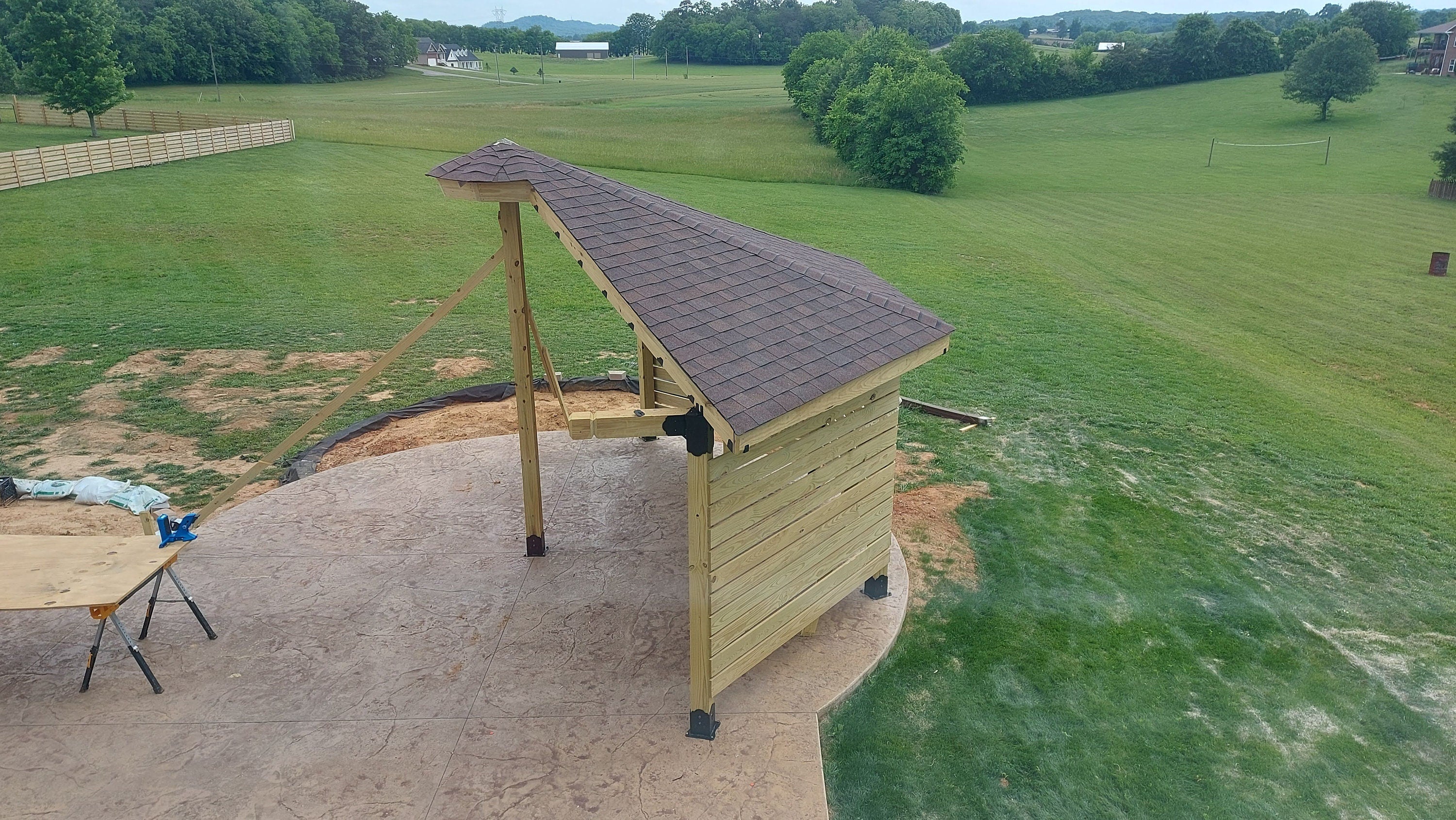 A wooden shelter with a sloped roof sits in a lush green field, surrounded by trees. The structure is newly built, emphasizing its sturdy design and fresh foundation amid a serene landscape.