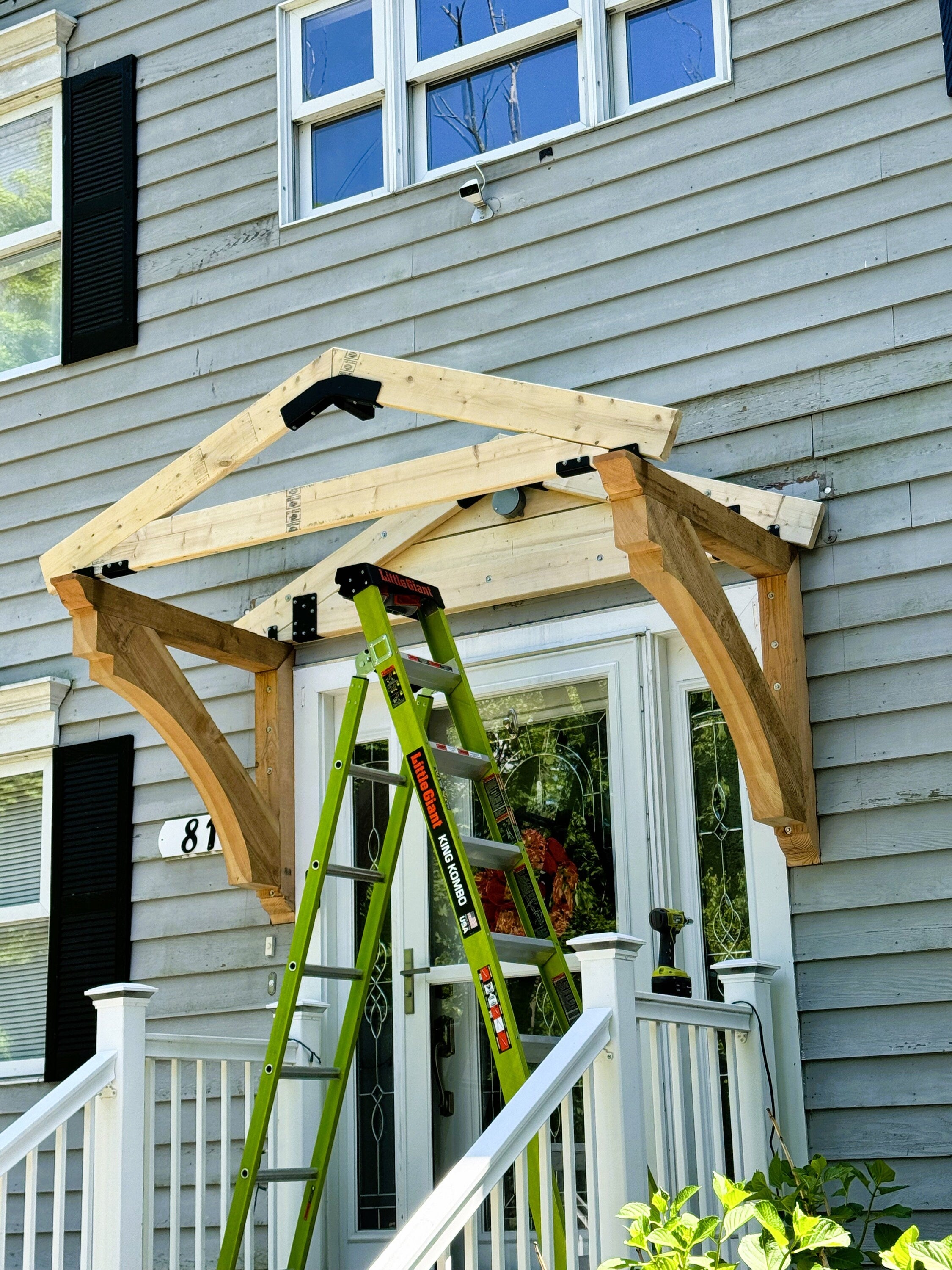 A worker uses a ladder to assemble a wooden roof structure above the front door of a house, highlighting craftsmanship and home improvement in progress.