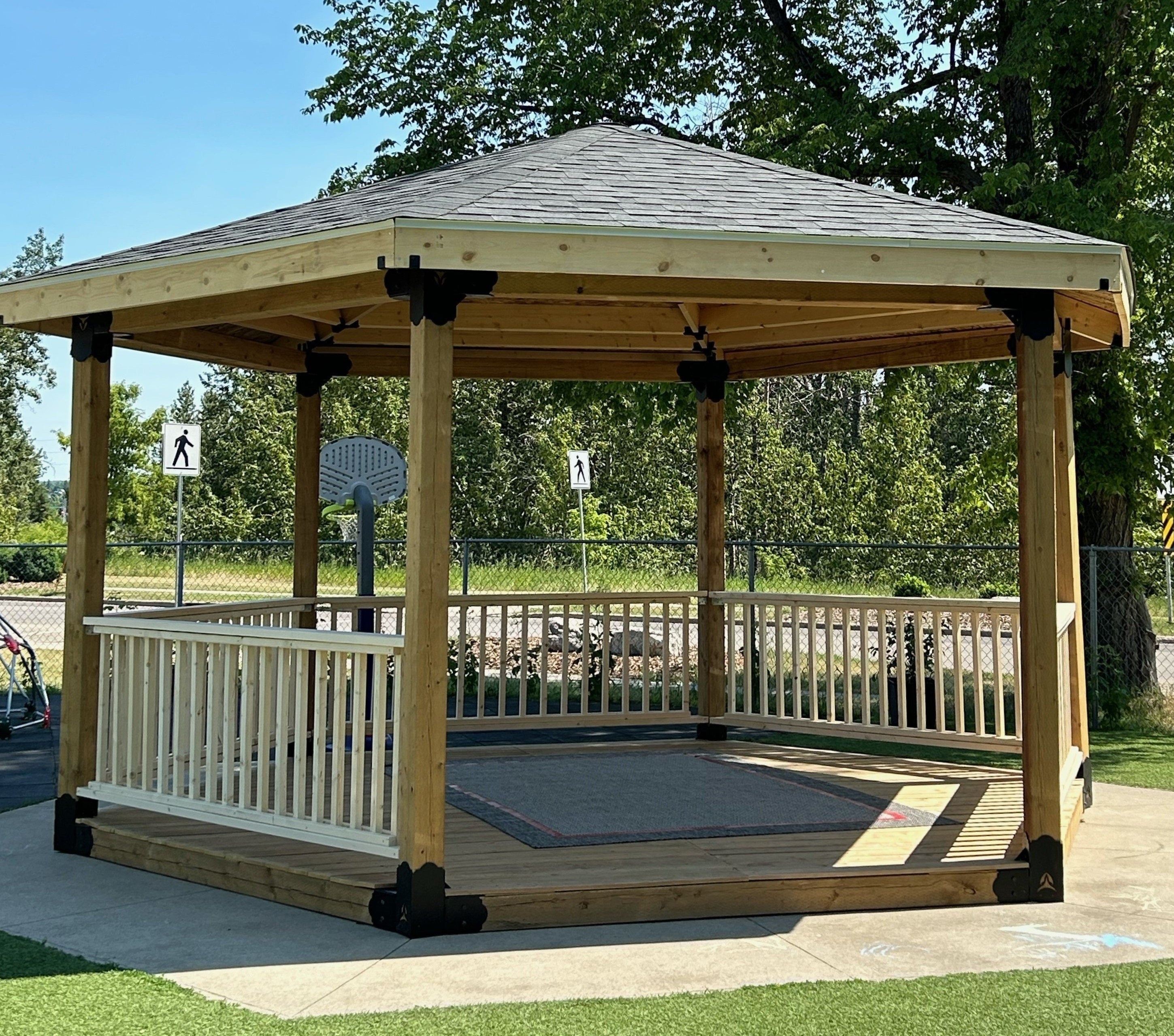 A wooden gazebo with a sloped roof stands in a sunny park, providing a shaded area for relaxation. Lush green grass surrounds the structure and nearby signs indicate pedestrian pathways.