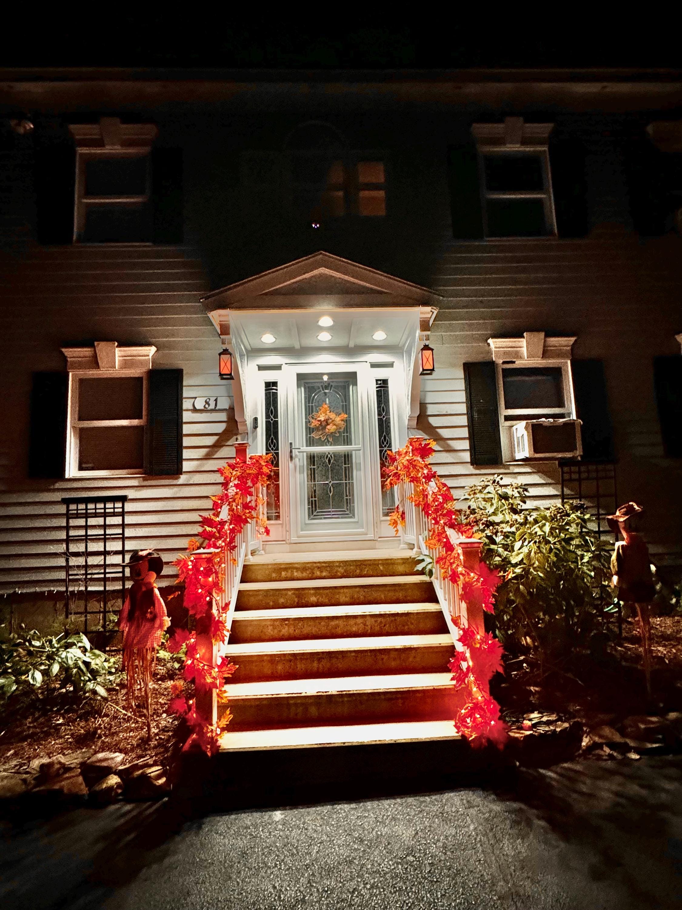 The entrance of the house is adorned with vibrant autumn leaves and glowing lanterns, creating a warm ambiance in the quiet night. It invites all who pass by.